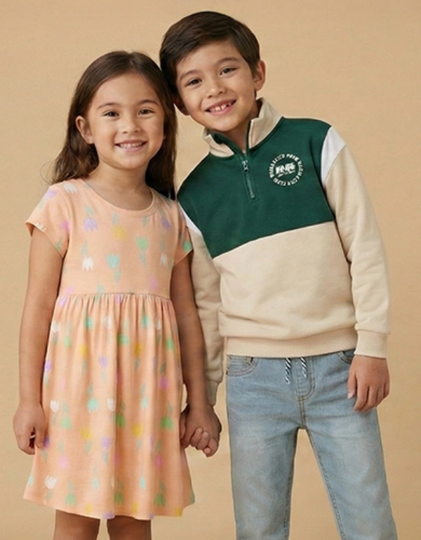 Dvě děti stojí společně před jednobarevným béžovým pozadím. Dívka má na sobě barevné šaty, zatímco chlapec má zelenou a krémovou mikinu.